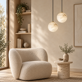 A modern beige boucle armchair in a warm, sunlit minimalist living room with a marble side table, hanging pendant lights, and a built-in wooden shelf.