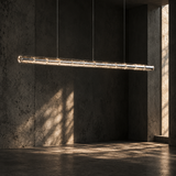 Illuminated clear glass linear chandelier suspended in a dark concrete room with sunlight falling across the wall.