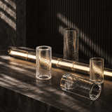 Linear chandelier resting on a dark stone surface beside separate textured glass cylinders, with its central light illuminated.