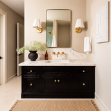 A styled bathroom vanity featuring a black cabinet with gold hardware, a white marble countertop, a rectangular mirror, and matching wall sconces. The scene includes a vase of greenery, a towel, and a woven rug in a warm neutral-toned bathroom.