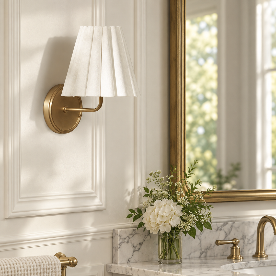 A wall-mounted sconce with a white pleated drum shade and brushed brass hardware, installed on a white paneled bathroom wall above a marble vanity with a gold-framed mirror and flowers.