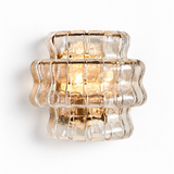 A modern wall sconce featuring a textured, ribbed clear glass shade with a warm gold metallic frame and glowing light inside, shown against a plain white background.