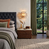 A modern bedroom scene featuring a glowing crystal chandelier wall sconce mounted on a beige wall above a wooden nightstand. The room includes a bed with gray bedding, green and rust accent pillows, olive green curtains, and a large window looking out to green foliage.