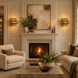 A beautifully styled living room featuring a central fireplace with a warm fire burning inside, flanked by two ornate wall sconces and a large abstract painting above. The room is furnished with cream-colored armchairs, a matching sofa, a marble coffee table with books and a plant, and a neutral beige paneled wall.