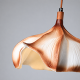 A close-up of a sculptural pendant light shade with a warm brown and cream marbled or petal-like surface, suspended against a plain light gray background.