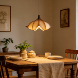 A warm, flower-shaped pendant light hangs over a rustic wooden dining table set with plates, a linen runner, and small ceramic plants in a cozy, warmly lit room.