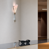 A modern wall-mounted sconce with two angled translucent shades and a polished metallic base, installed on a beige wall above a reflective dark counter. Black sunglasses rest on the counter, and an interior hallway is visible in the background.