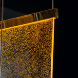 Close-up of the staircase chandelier's golden acrylic panel glowing with tiny bubbles, hung by slim cables against a dark background.