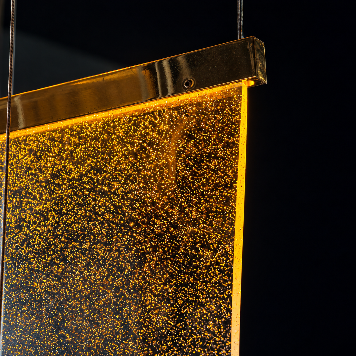 Close-up of the staircase chandelier's golden acrylic panel glowing with tiny bubbles, hung by slim cables against a dark background.