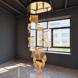 A modern multi-tiered chandelier with wavy, translucent glass panels and warm gold-toned metal accents, suspended from a circular ceiling fixture in a minimalist room with dark gray walls and a large window.