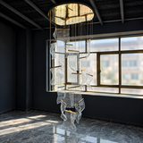 A large modern chandelier with a circular gold-toned top canopy and cascading clear glass or acrylic wave-like panels suspended by thin rods, hanging in a dark interior room with large windows and a marble floor.