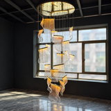 A large modern chandelier with multiple cascading, translucent wave-like glass panels illuminated from within, hanging in a spacious dark room with a large window and polished floor.