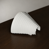 White steel wall sconce resting on a dark wooden surface, showing its curved overlapping slats from an angled side view.