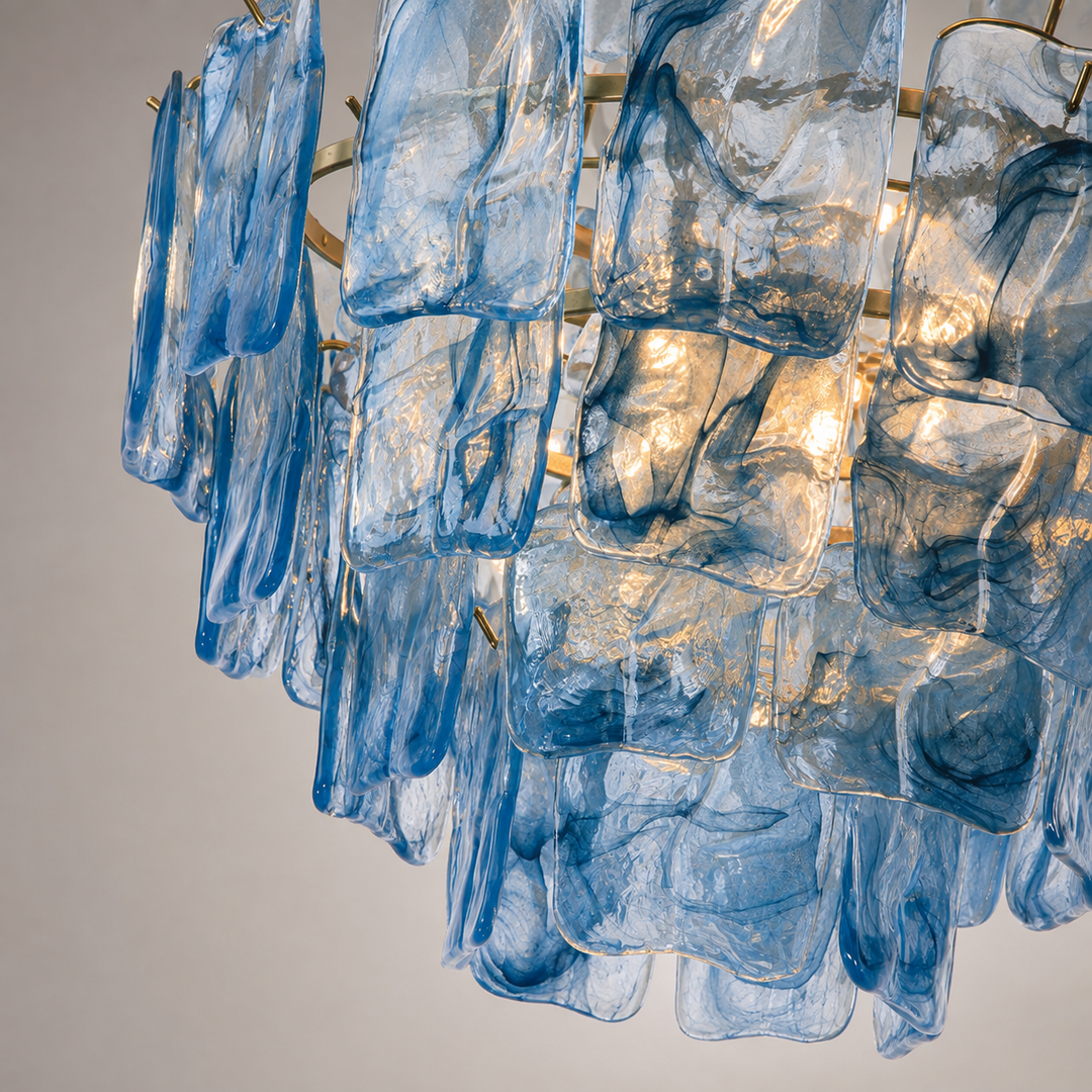 A close-up of a modern chandelier featuring multiple curved, translucent blue glass panels suspended from a gold-toned metal frame against a neutral light gray background.