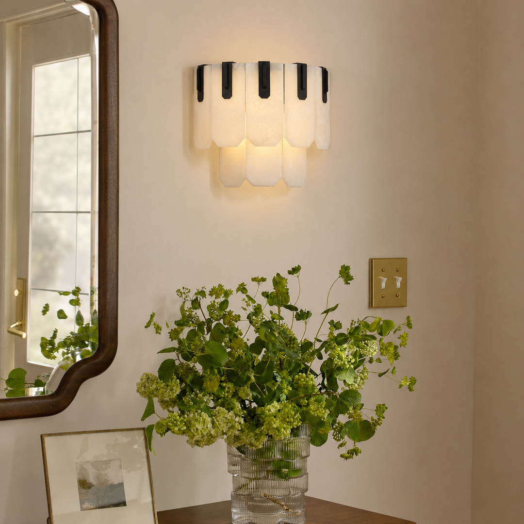 A modern wall sconce with white scalloped glass shades and black vertical accents, mounted on a beige wall above a table with a green floral arrangement.