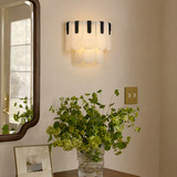 A modern wall sconce with white scalloped glass shades and black vertical accents, mounted on a beige wall above a table with a green floral arrangement.