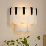 Wall sconce with layered white stone panels and black iron brackets casts a warm glow on a beige wall above greenery.