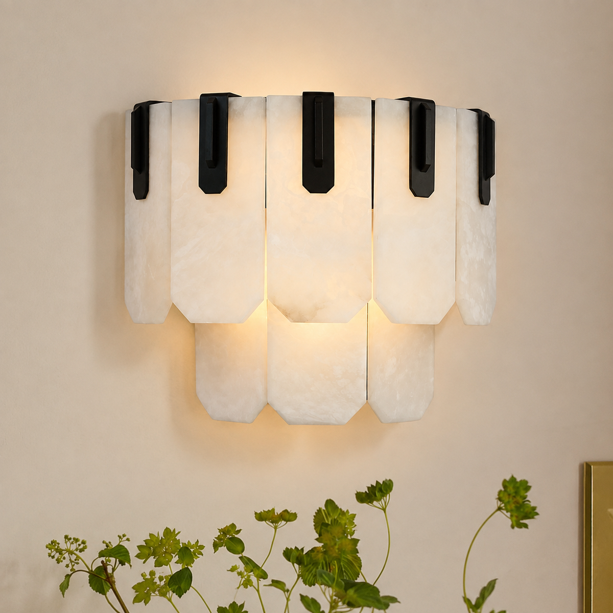Wall sconce with layered white stone panels and black iron brackets casts a warm glow on a beige wall above greenery.