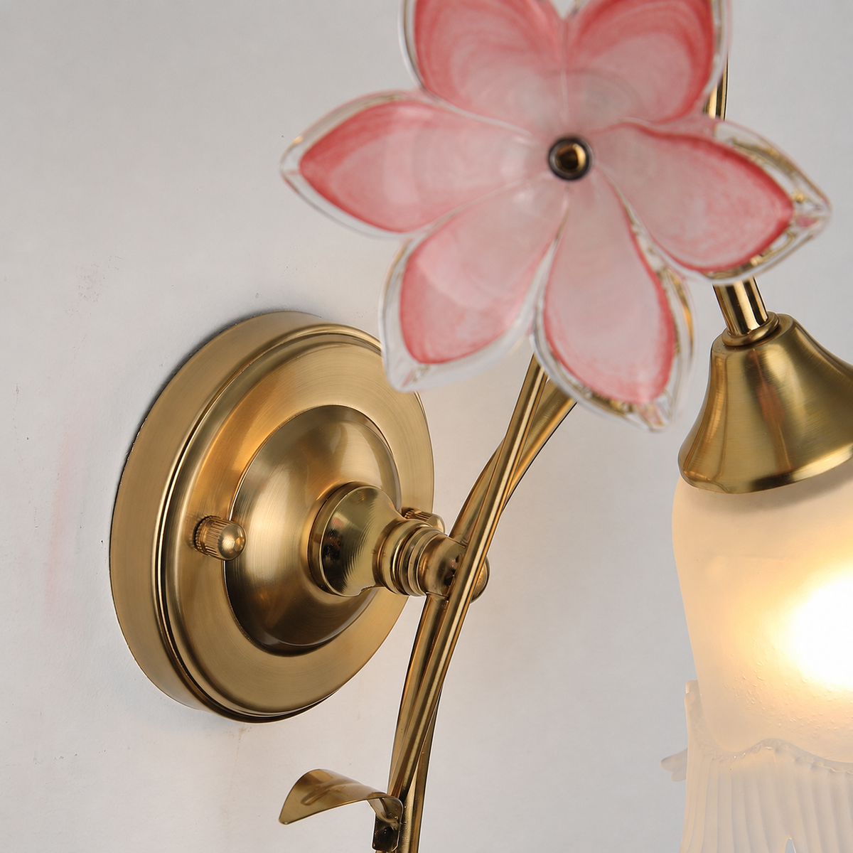 Close-up of a gold brass wall sconce with a pink glass flower and glowing frosted bell shade on a pale wall.