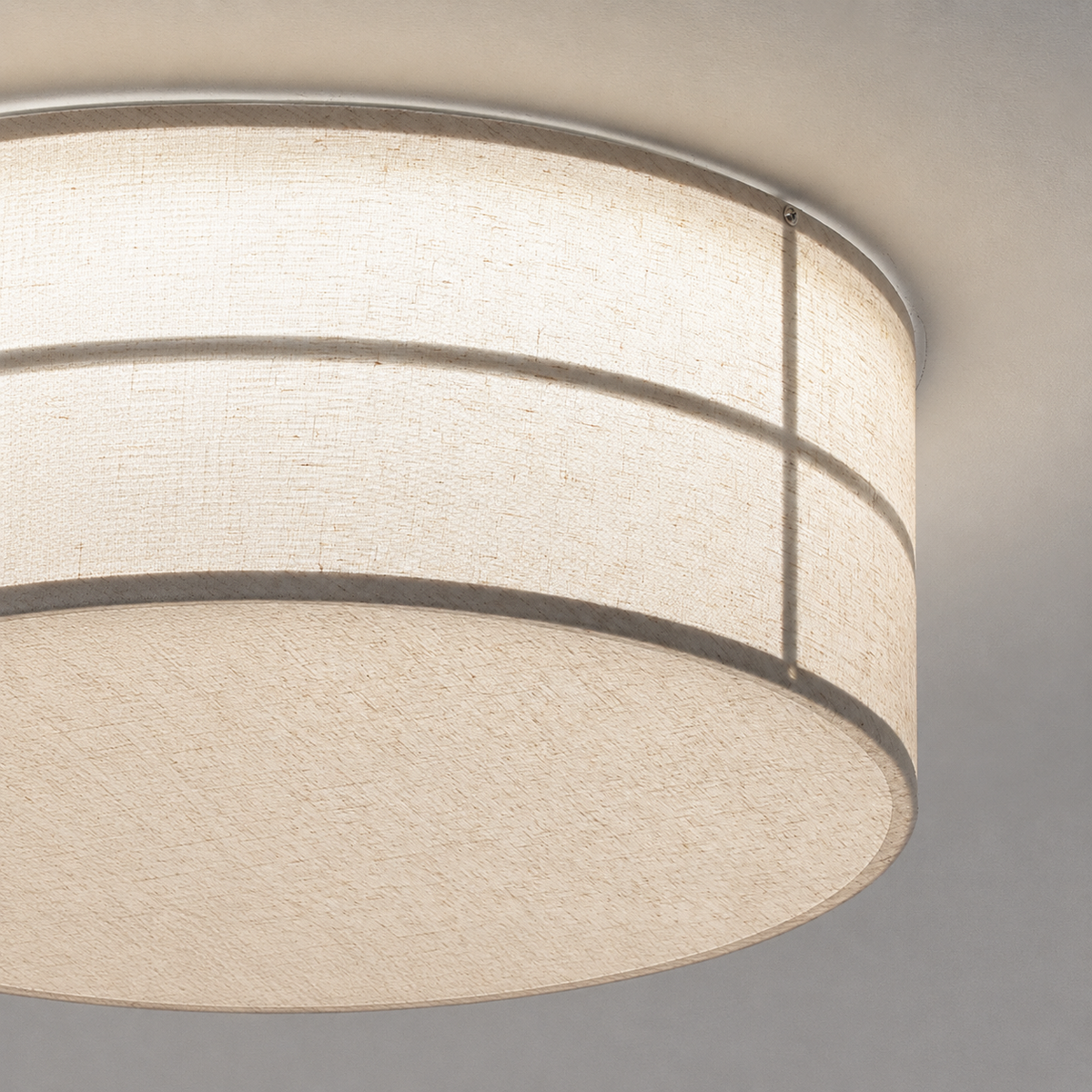 A close-up of a cylindrical ceiling light fixture with a textured cream fabric drum shade and a thin metallic trim, mounted against a neutral gray ceiling.