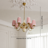 A six-light chandelier with gold-toned metal arms and cylindrical shades featuring a pink and white geometric pattern, shown hanging in a white interior room. Measurement overlays indicate a diameter of 83 cm (32.