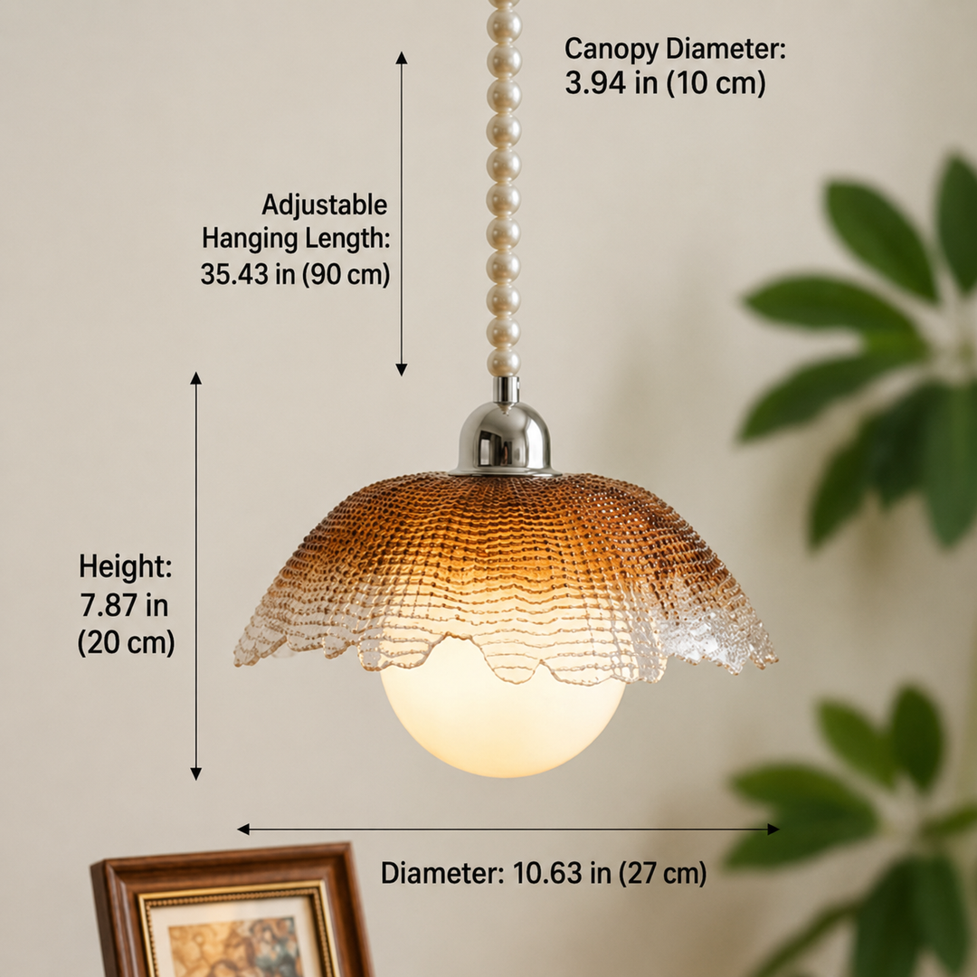 A pendant light shade with overlaid dimension labels showing canopy diameter, hanging length, height, and overall diameter. The lamp has a textured amber-brown dome shade, a glowing white globe bulb, a silver metal top, and a pearl-bead hanging cord against a beige wall with blurred green leaves and a framed picture in the background.