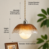 A pendant light shade with overlaid dimension labels showing canopy diameter, hanging length, height, and overall diameter. The lamp has a textured amber-brown dome shade, a glowing white globe bulb, a silver metal top, and a pearl-bead hanging cord against a beige wall with blurred green leaves and a framed picture in the background.