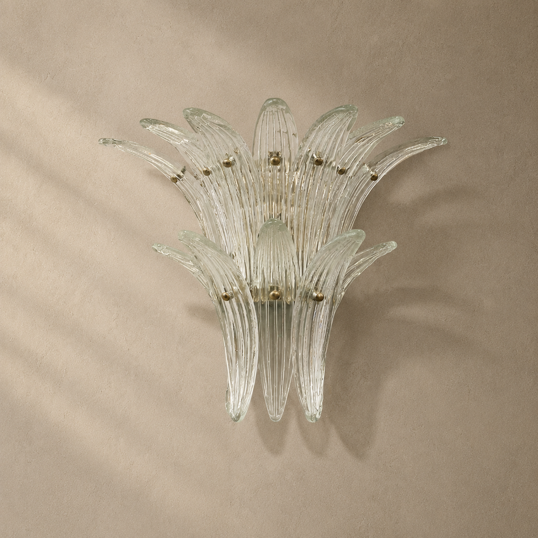 A clear ribbed glass wall sconce with multiple curved petal-like arms, mounted on a warm beige textured wall with soft shadows.