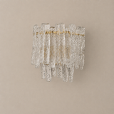 Wall sconce of hand-textured clear-to-amber glass pieces on a brass-tone frame, unlit on a plain beige wall.