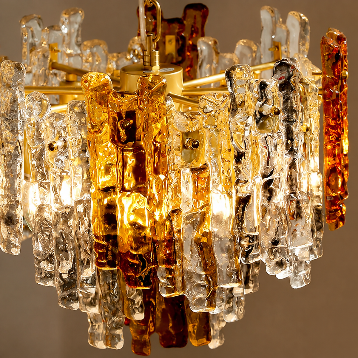 A close-up of a mid-century modern chandelier featuring textured amber and clear glass rods hanging from a brass-toned metal frame, illuminated with warm light against a neutral background.