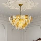 A modern chandelier with irregular, organic-shaped translucent amber and clear glass or acrylic pieces, suspended by a gold chain from an ornate white decorative ceiling medallion in a classic room interior.