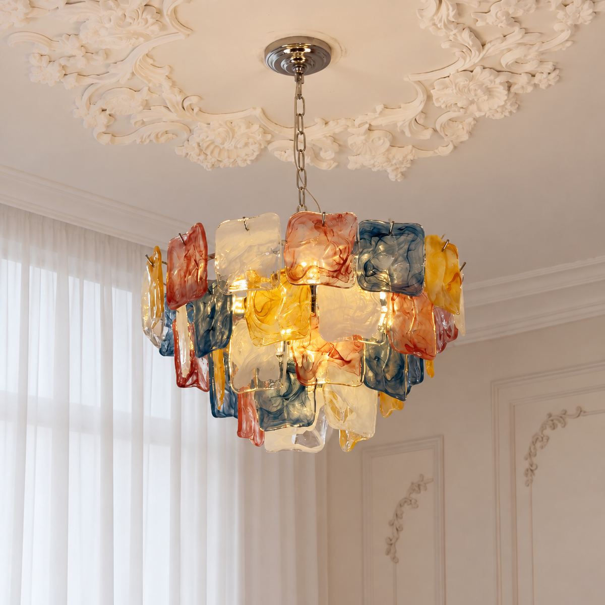 A colorful glass chandelier with multiple rectangular glass panels in red, blue, yellow, and clear tones, hanging from a chain in an ornate cream-colored room with decorative ceiling molding.