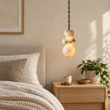 A single-light marble chandelier on a black chain glows above a wooden bedside table beside a neutral upholstered bed.