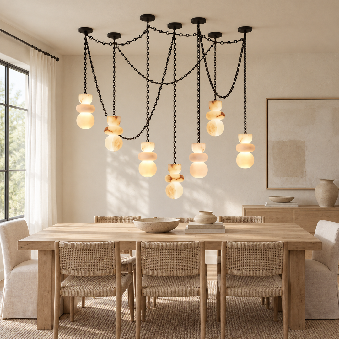 A modern multi-light pendant chandelier with black chain structures and stacked amber glass globes, hanging over a light wood dining table with woven chairs in a bright, neutral-toned dining room.