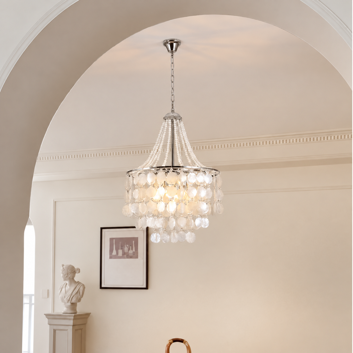 A multi-tiered crystal chandelier with a silver-toned metal frame and hanging beads, suspended from the ceiling in a bright, elegant room with an arched doorway and framed wall art.