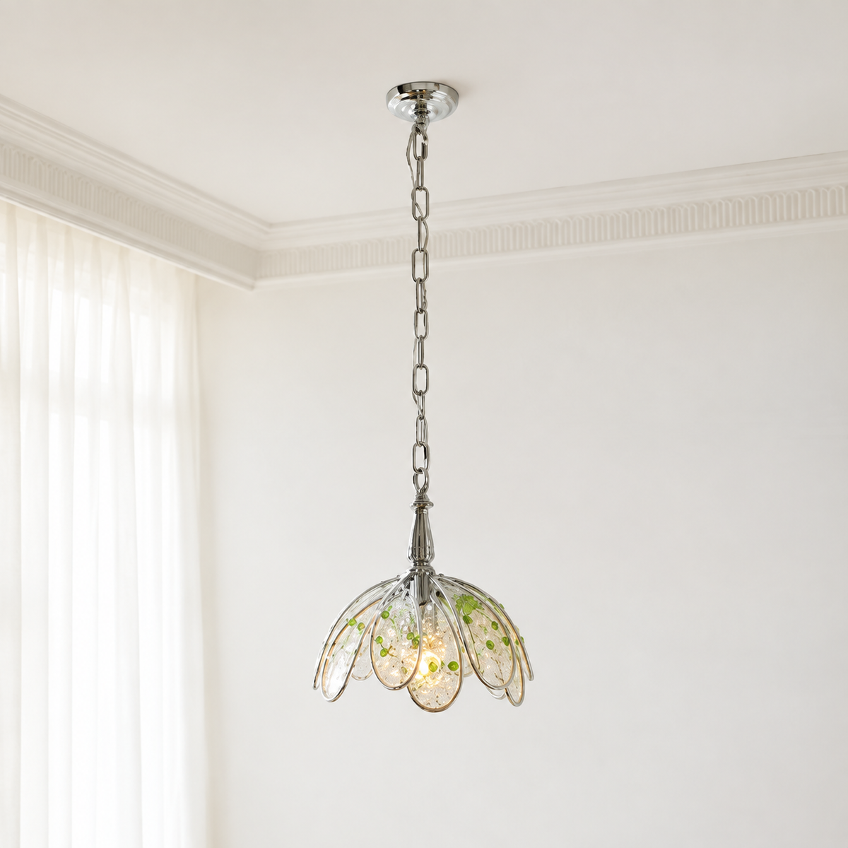 Clear glass petal chandelier with green floral accents hangs from a silver-toned chain in a white room beside sheer curtains.
