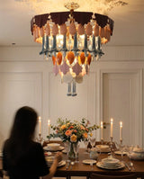 A large, ornate chandelier with layered amber, peach, and blue glass leaf-like elements hangs over a beautifully set dining table with candles and a floral centerpiece. A woman is seated at the table in a warmly lit dining room.