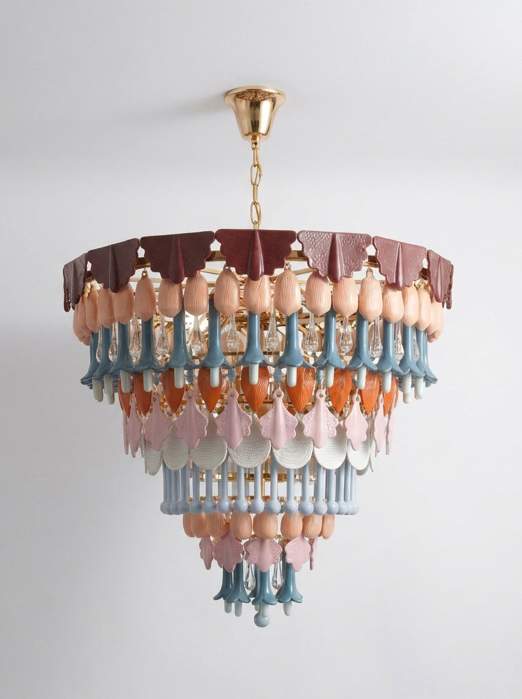 A multi-tiered chandelier with a gold metal frame and canopy, decorated with layers of dusty pink, light blue, and terracotta-colored sculptural beads and leaf-shaped drops, hanging against a plain light gray wall.