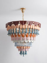 A multi-tiered chandelier with a gold metal frame and canopy, decorated with layers of dusty pink, light blue, and terracotta-colored sculptural beads and leaf-shaped drops, hanging against a plain light gray wall.
