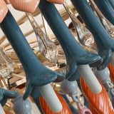 Close-up of a decorative chandelier with glossy teal-blue floral-shaped metal arms, clear glass teardrop pendants, white cylindrical accents, and orange ribbed hanging elements.