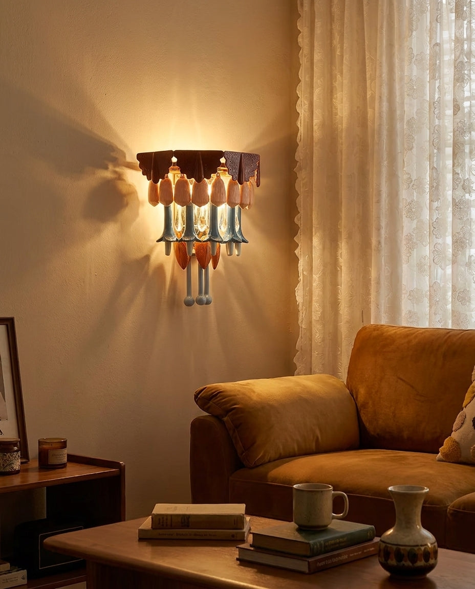 Chandelier with multicolor glass drops glows against a beige living room wall beside a mustard armchair and sheer curtains.