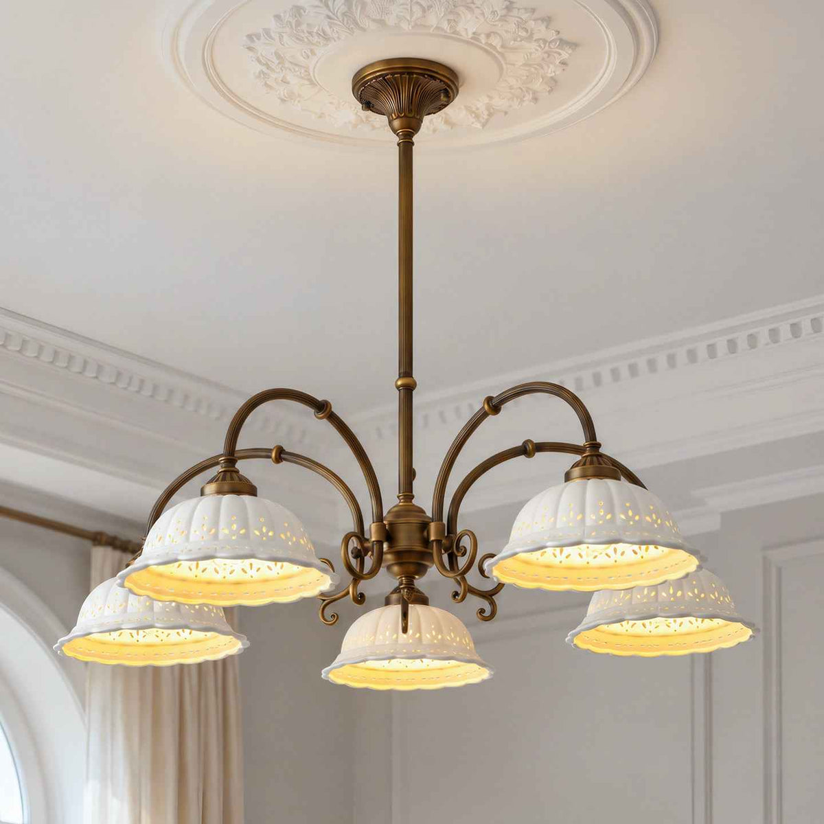 An ornate five-light chandelier with a bronze-finished metal frame and white lace-detailed bell shades, illuminated in a classic European-style room with white walls and molding.