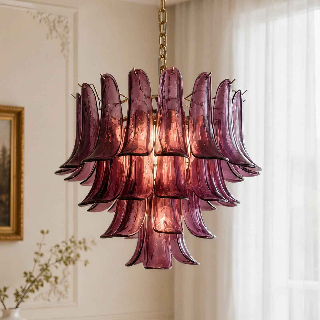 A modern chandelier with multiple layered, curved purple glass shades hanging from a gold chain in a bright, elegant room with white walls and sheer curtains.