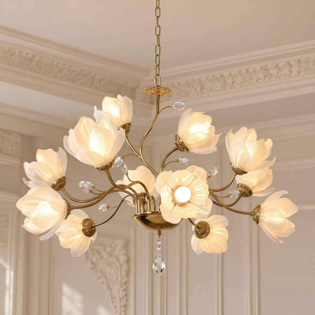 A brass chandelier with 12 flower-shaped glass shades and hanging crystal prisms, shown lit in an elegant cream-colored room with decorative molding.