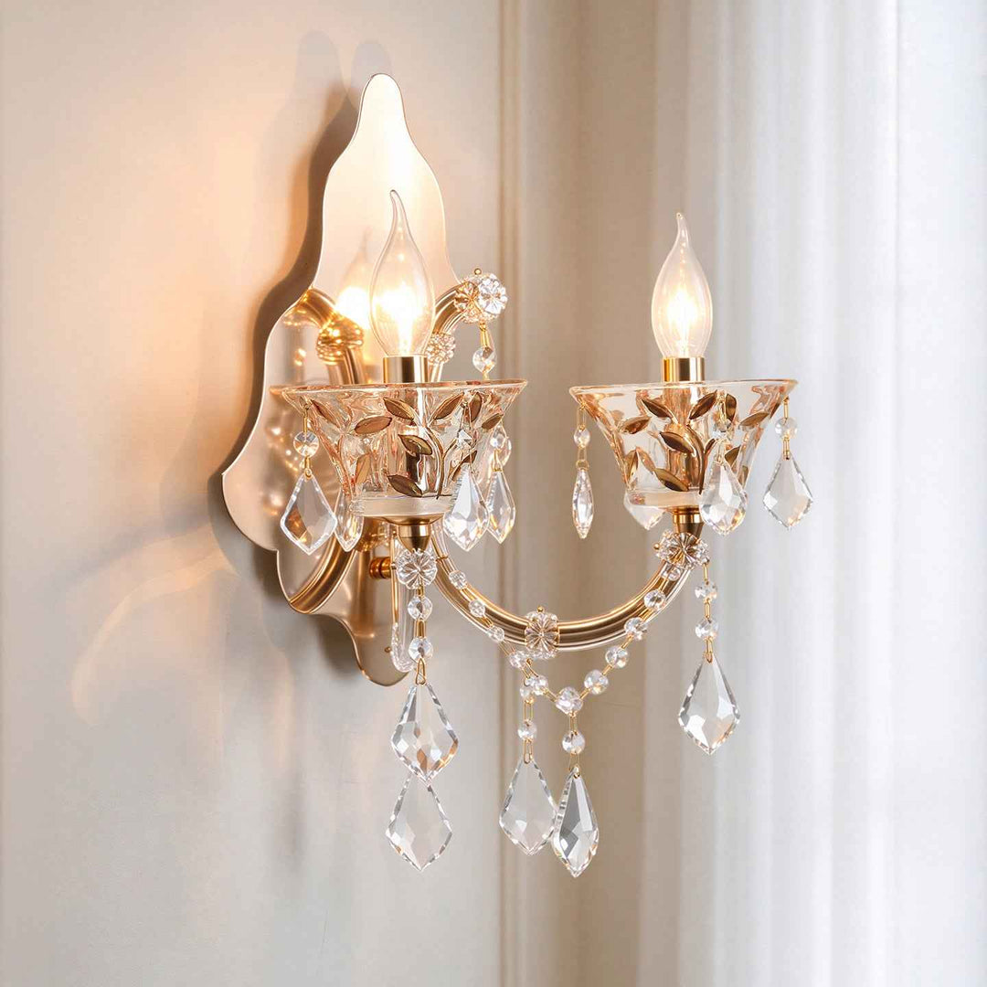 A two-light wall sconce with a warm gold/brass finish, featuring clear crystal teardrop pendants and candle-style bulbs glowing against a soft beige wall and curtain background.