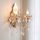 A two-light wall sconce with a warm gold/brass finish, featuring clear crystal teardrop pendants and candle-style bulbs glowing against a soft beige wall and curtain background.