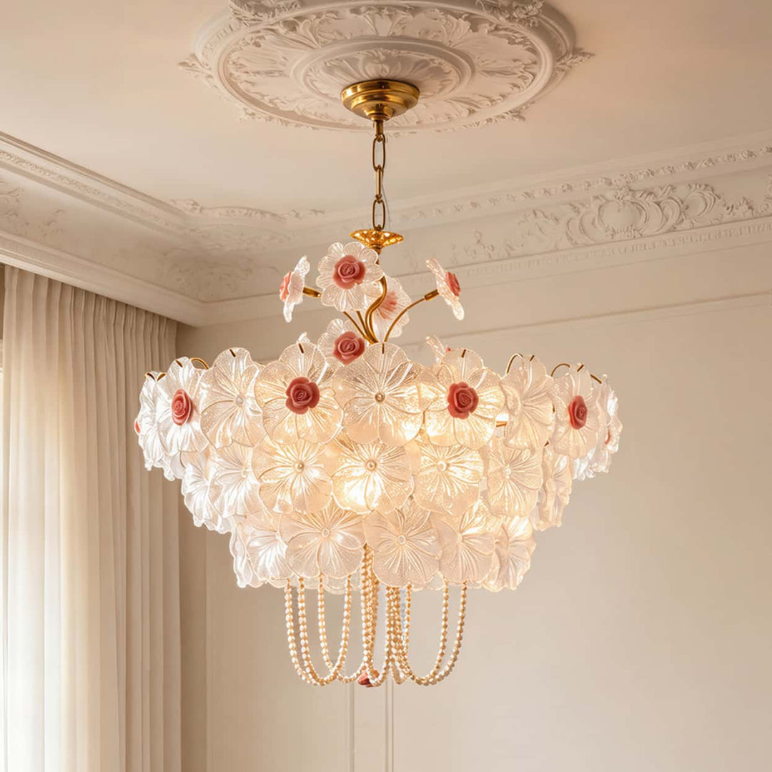 A large multi-tiered floral chandelier with white glass flower petals, pink rose accents, and hanging pearl-like strands, suspended from a decorative gold ceiling canopy in an ornate cream-colored room.