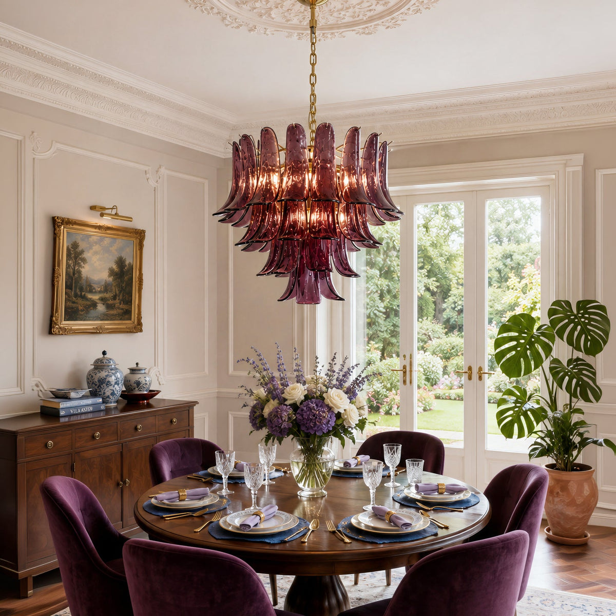 A large, elegant chandelier with layered, fluted glass shades in a deep plum purple hangs over a round wooden dining table set for six in a classic dining room. The room features cream paneled walls, a dark wood sideboard, and a large potted monstera plant in the background.