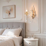 A gold wall sconce with hanging crystal prisms is mounted on a decorative cream paneled wall next to a bed with white linens and a gray upholstered headboard.