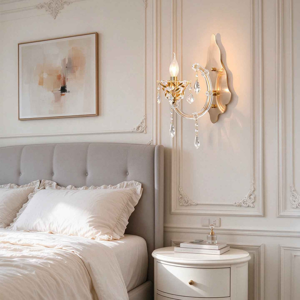 A gold wall sconce with hanging crystal prisms is mounted on a decorative cream paneled wall next to a bed with white linens and a gray upholstered headboard.